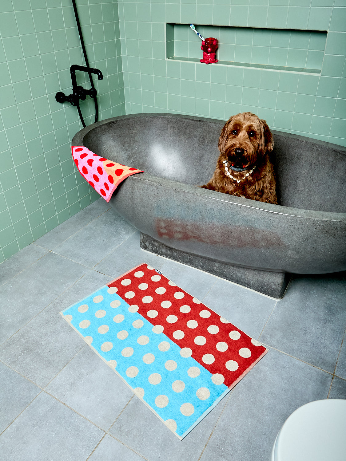 Dog in a bathtub with polka dot towels on a tiled bathroom floor.