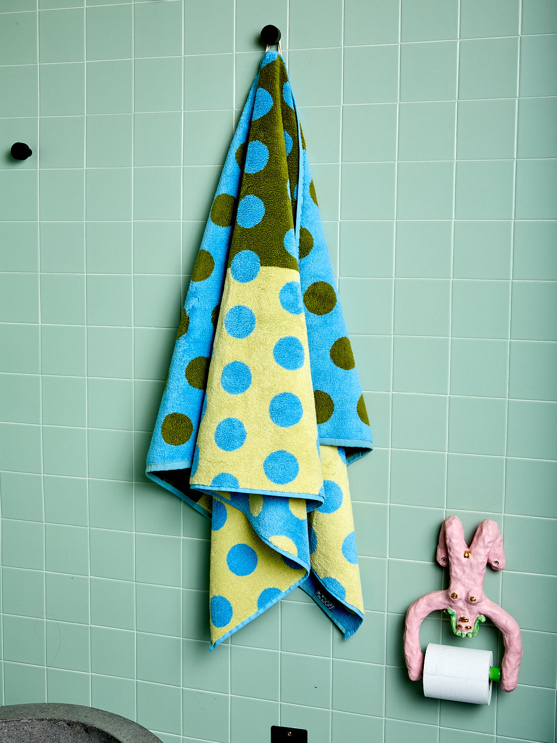 One green and yellow bath towel with blue polka dots hanging on a tiled wall next to a pink toilet paper holder.