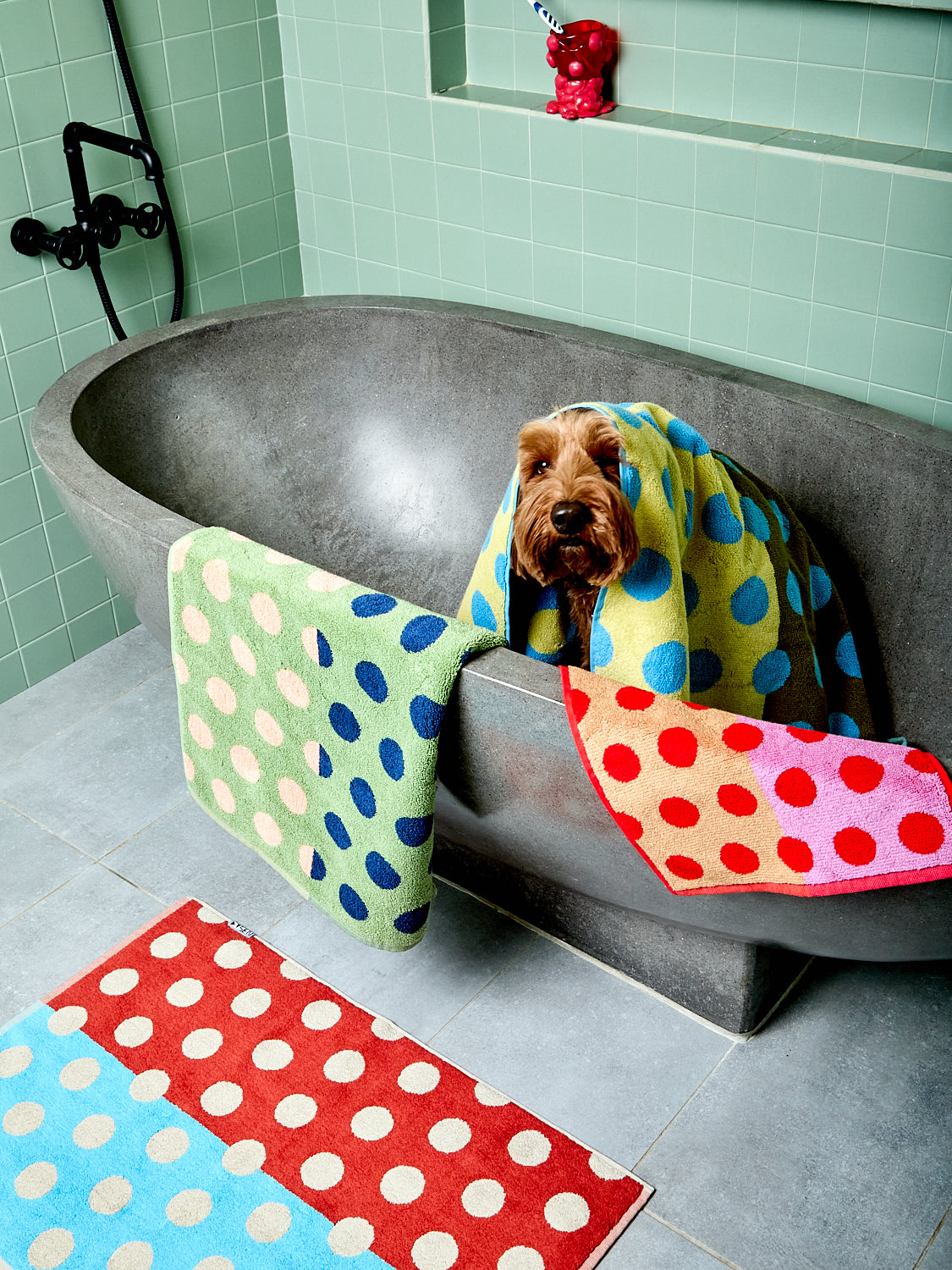 Dog wrapped in a colorful polka dot towel in a bathroom with tiled walls and floor.