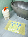 Light green bath mat featuring the word 'GEMINI' and an abstract two-sided face on a tile floor