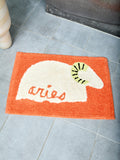 Orange bathmat with ivory bighorn sheep and text that reads "aries" on a tile floor
