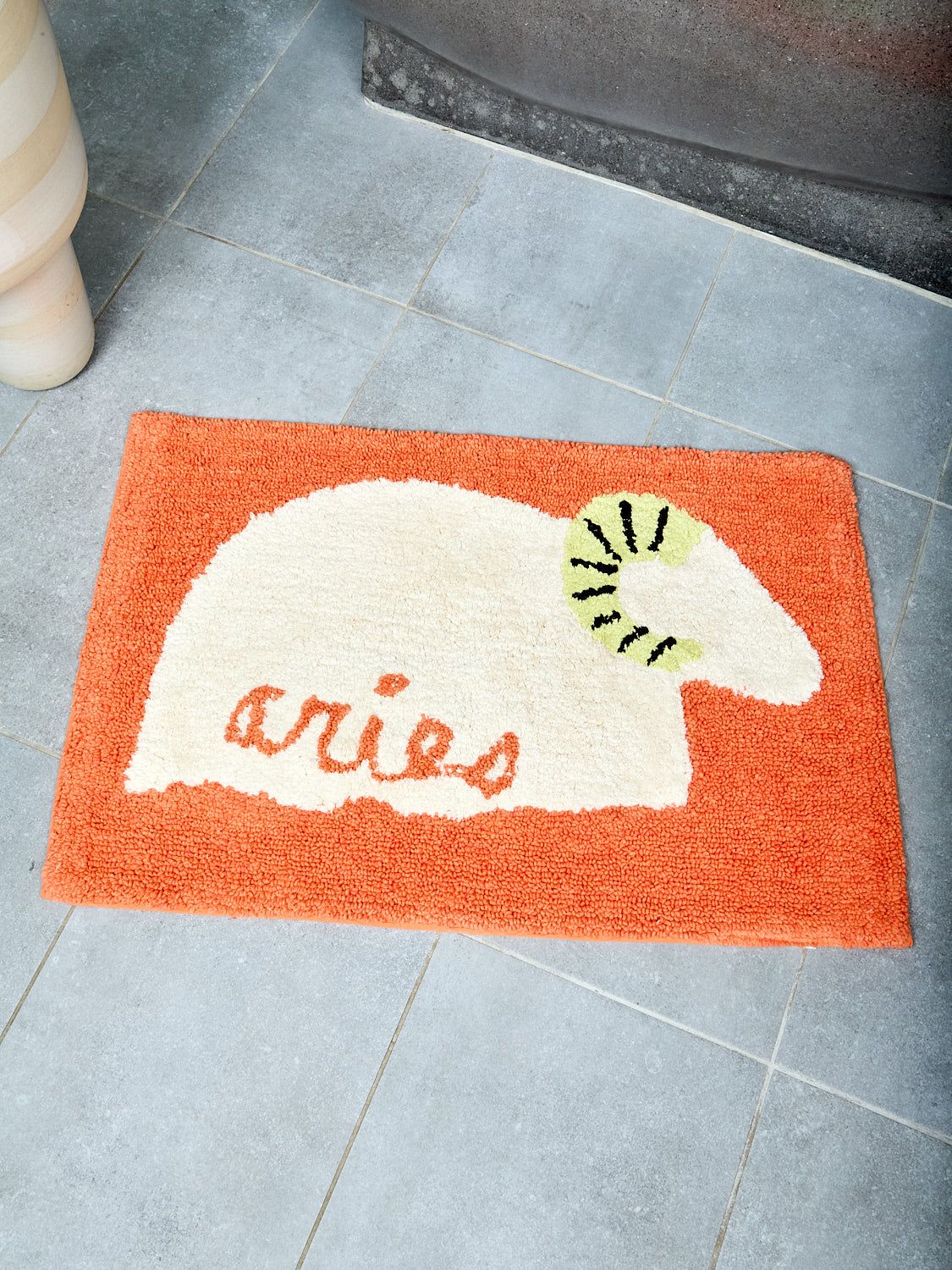 Orange bathmat with ivory bighorn sheep and text that reads "aries" on a tile floor