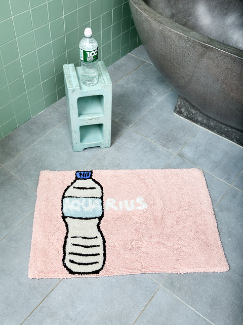 Pink bath mat with water bottle design and text reading "AQUARIUS" on a bathroom floor.
