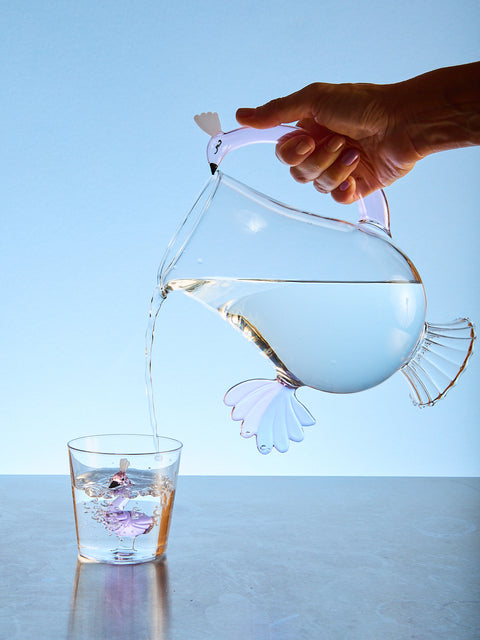 Glass pitcher with bird-shaped spout pouring water into a glass against a light blue background