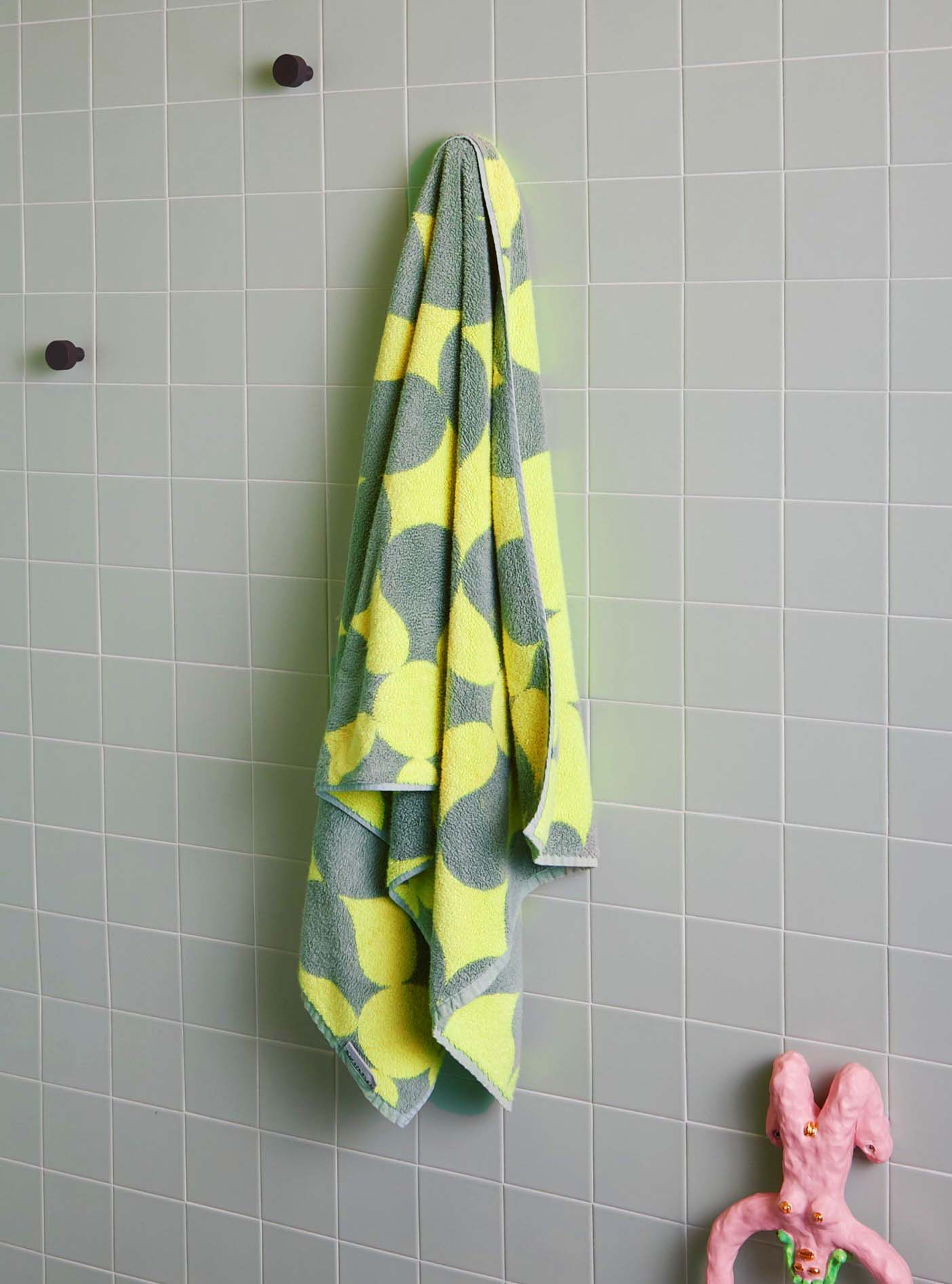 A sage green and light yellow patterned bath towel by Dusen Dusen hung on a light sage tiled wall.