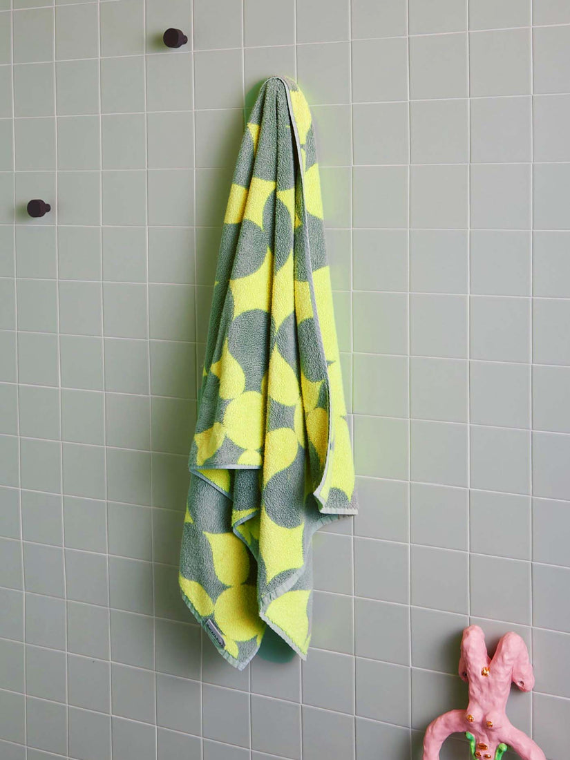 A sage green and light yellow patterned bath towel by Dusen Dusen hung on a light sage tiled wall.