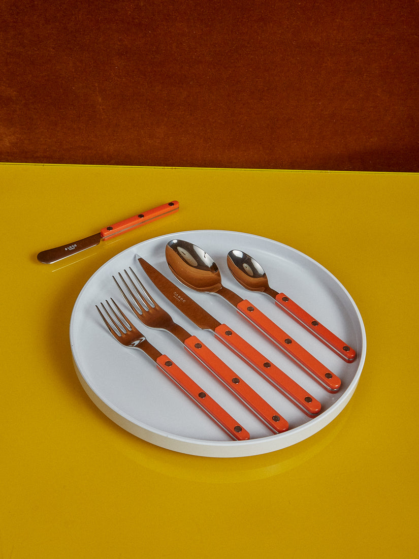 A white plate holds a orange bistro salad fork, dinner fork, dinner knife, dinner spoon, and teaspoon by Sabre. A spreader sits just above the plate.