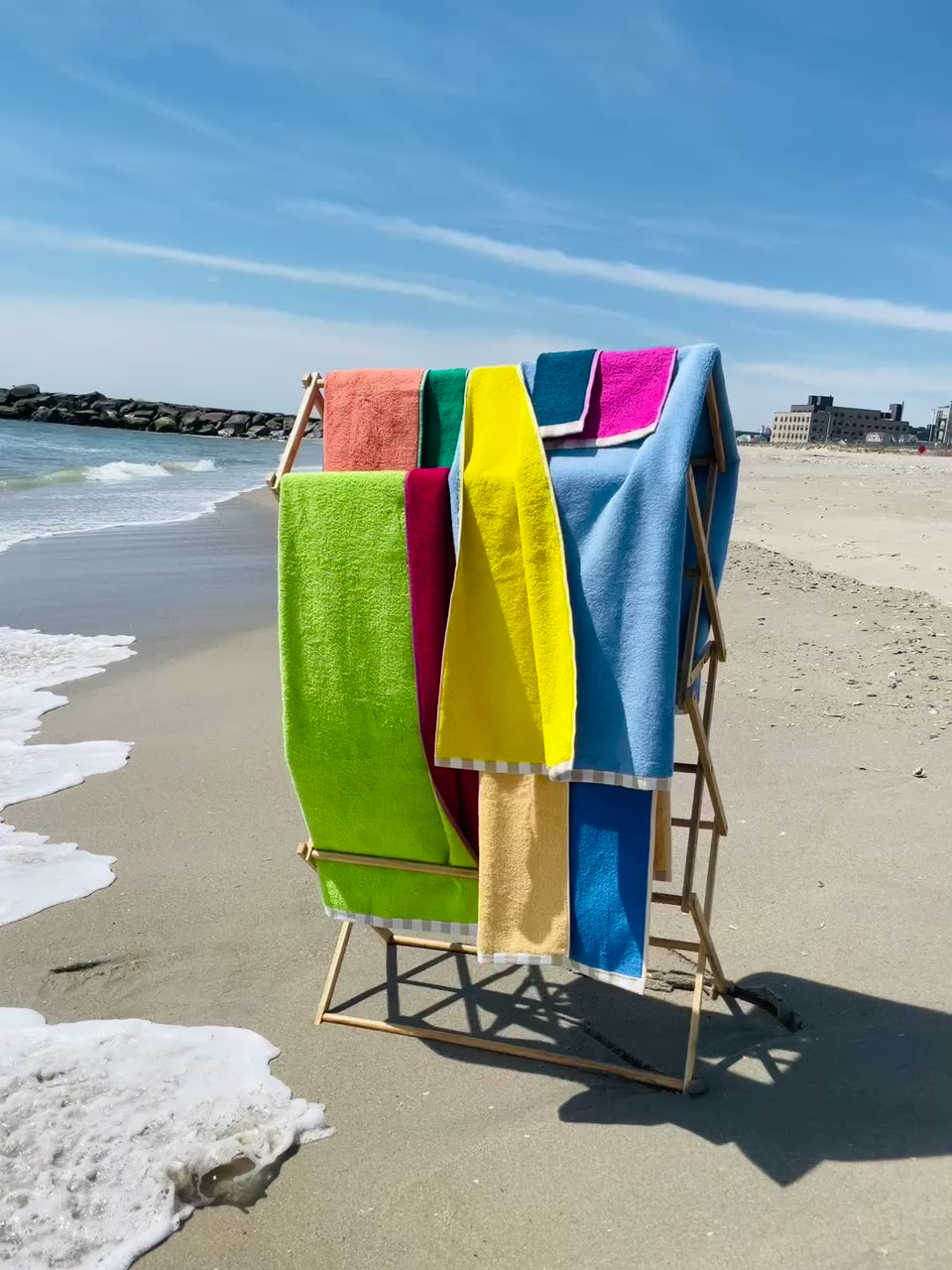 A drying rack full of reversible bath and hand towels sits at the shoreline of a beach with a subtle gust of wind.