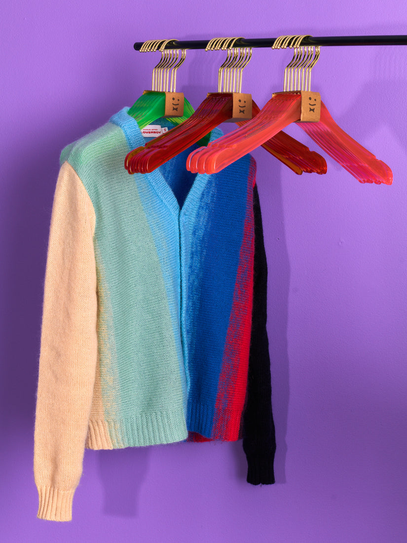 A clothing rack holds a green, orange, and pink set of a hangers. A multicolored cardigan hangs on the green set of hangers.