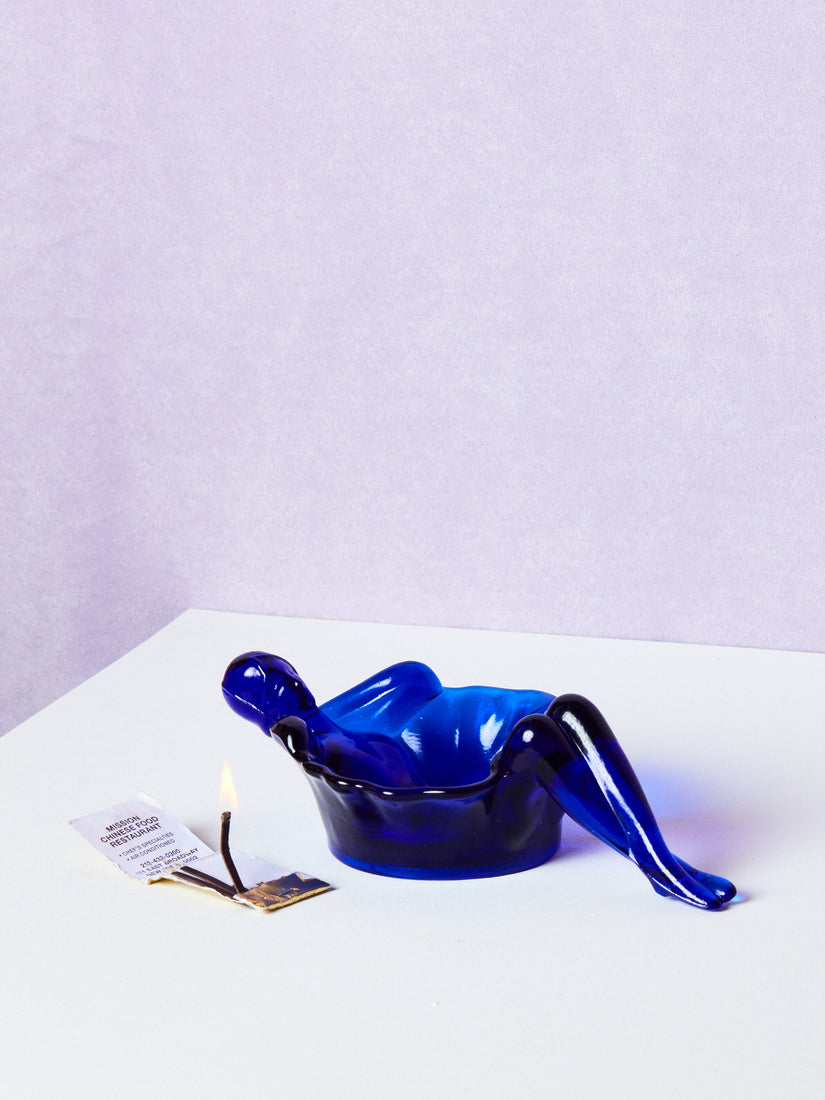 A cobalt blue bathing lady dish next to a lit book of matches.