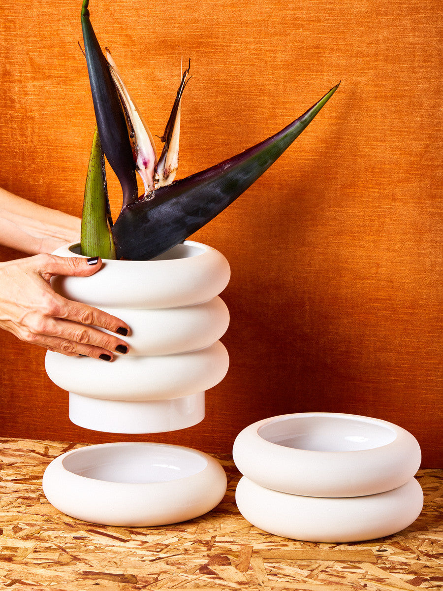 Hands lift up the tall white stacking planter from its saucer. A plant is potted inside the planter, and the short version sits right.