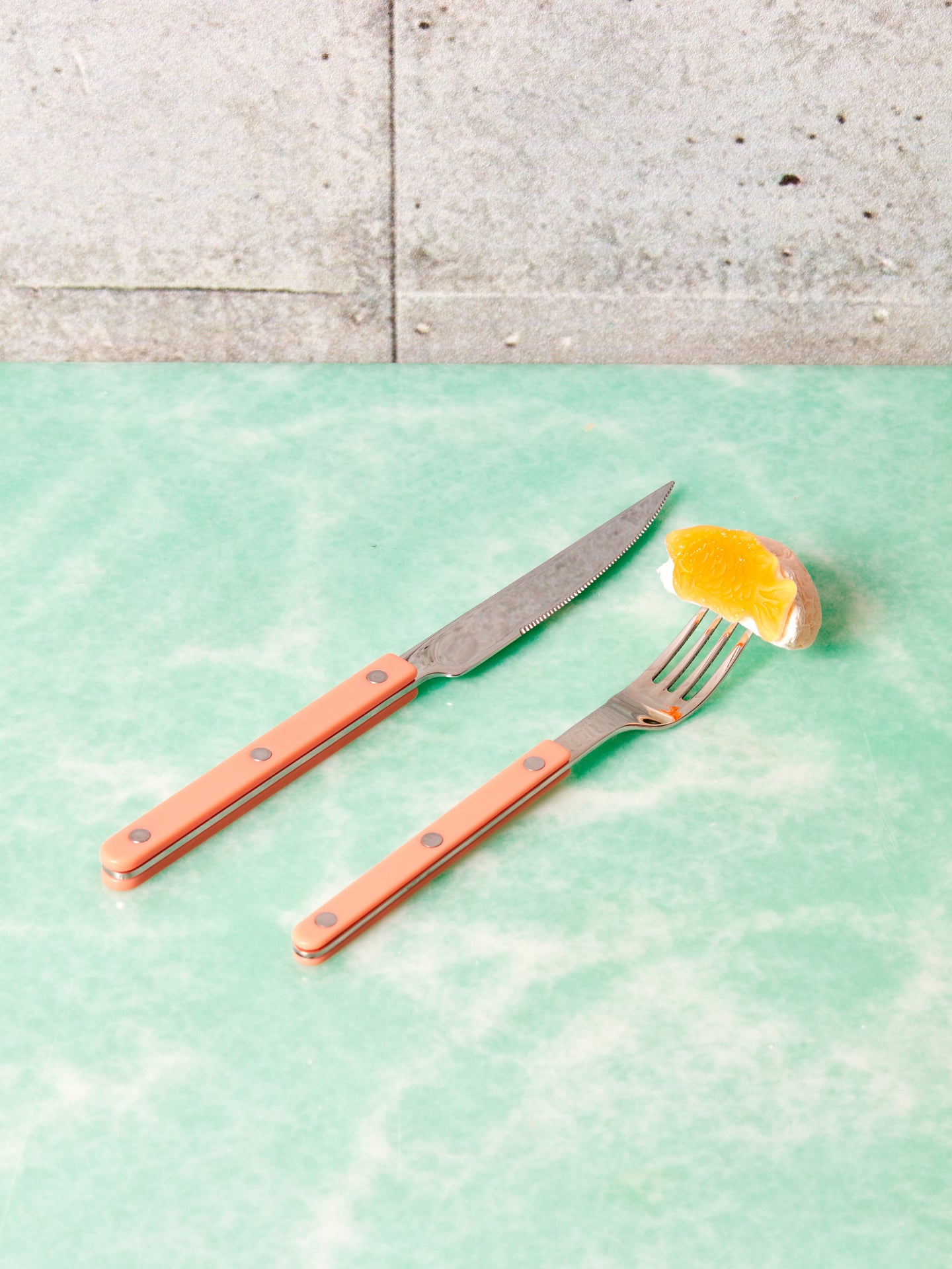 A coral sabre dinner knife next to a salad fork that has an orange slice on its prongs.