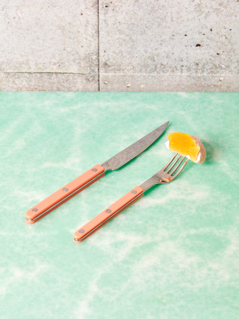 A coral sabre dinner knife next to a salad fork that has an orange slice on its prongs.