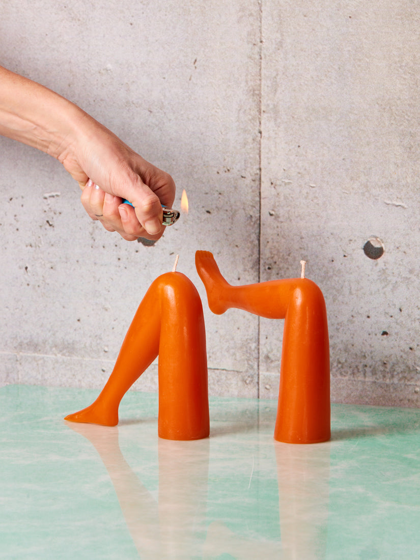 A pair of decorative orange candles in the shape of legs, one with its foot elevated, the other resting - being lit by a hand carrying a lighter.