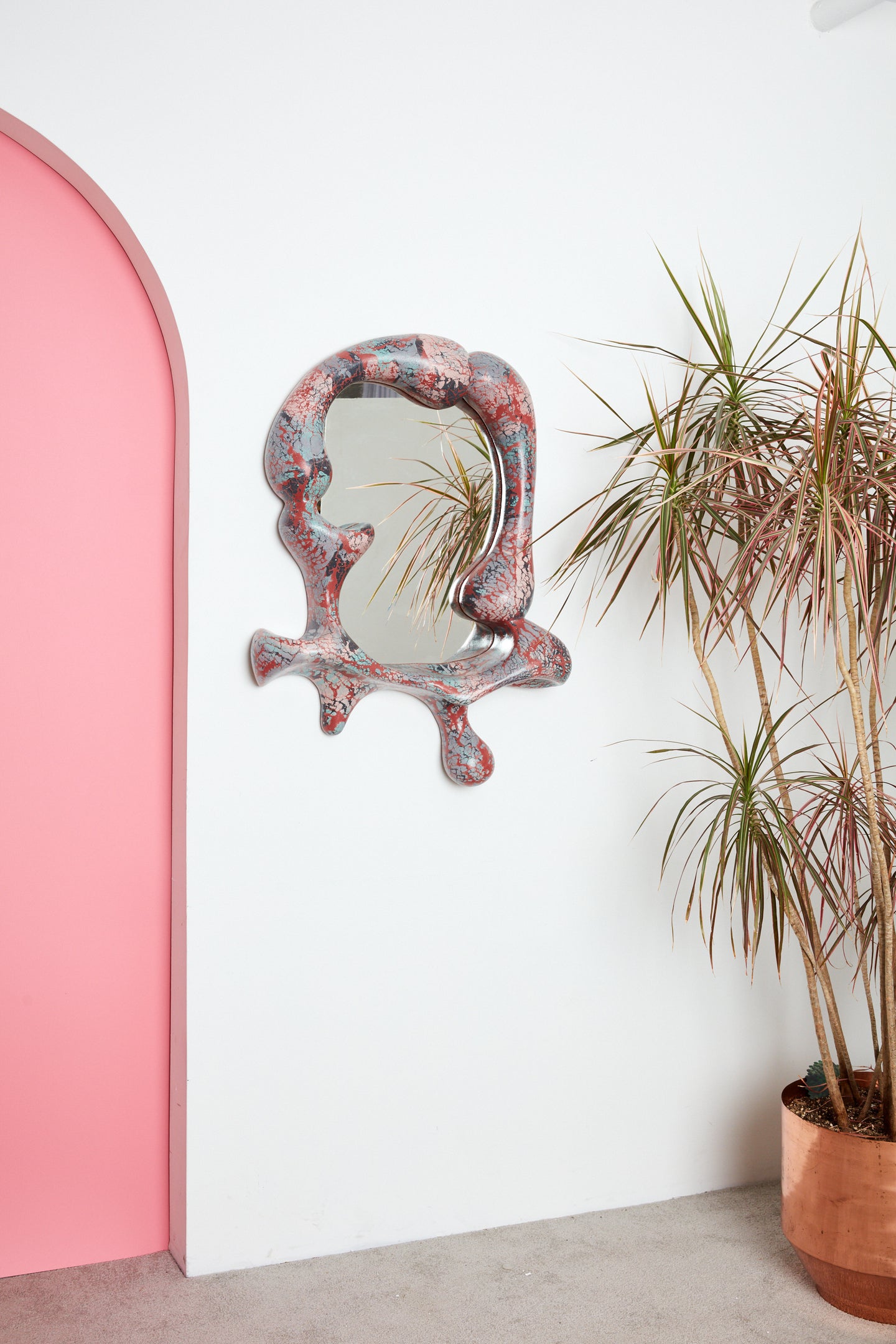 Iris Mirror by Concrete Cat hung on a white wall near a pink archway and a copper planter with a palm in it.