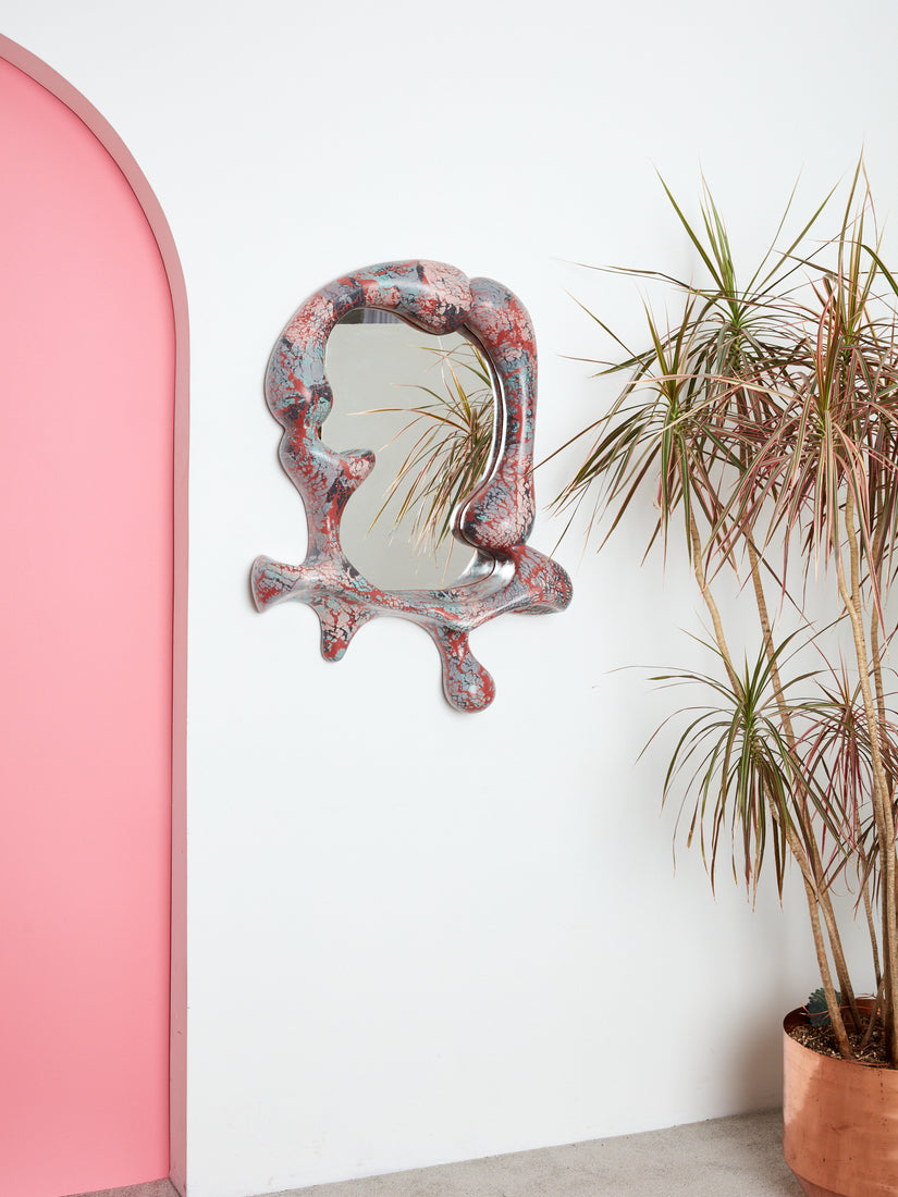 Iris Mirror by Concrete Cat hung on a white wall near a pink archway and a copper planter with a palm in it.