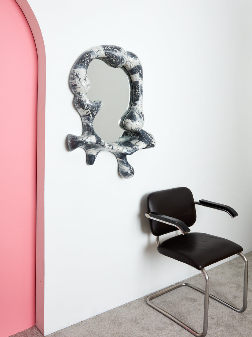 Kafka Iris Mirror by Concrete Cat hung on a wall above a vintage black chair.