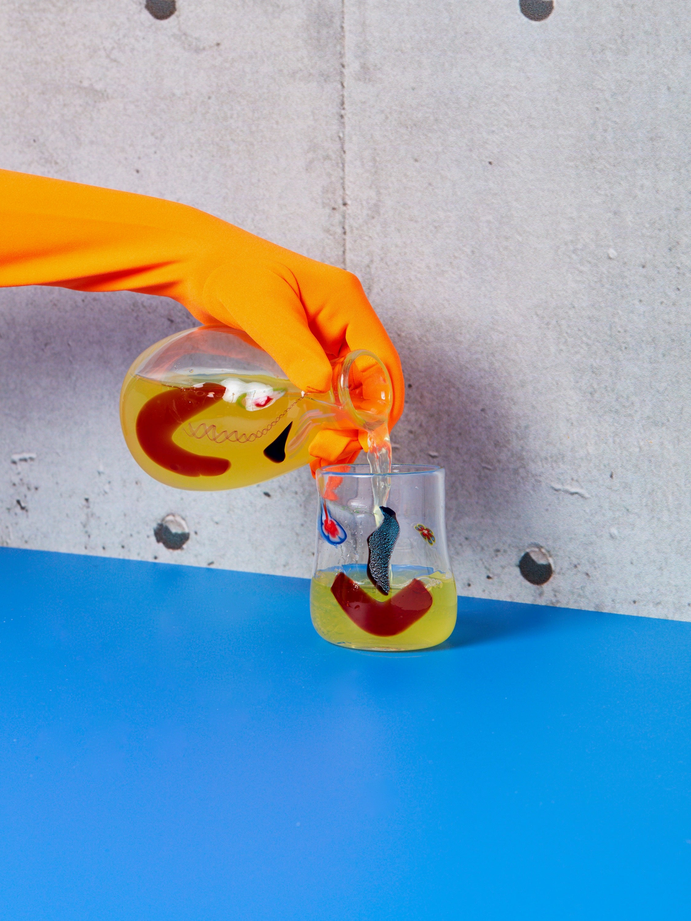 An orange gloved hand pours a yellow drink from a Face Carafe into a Face Vessel glass.