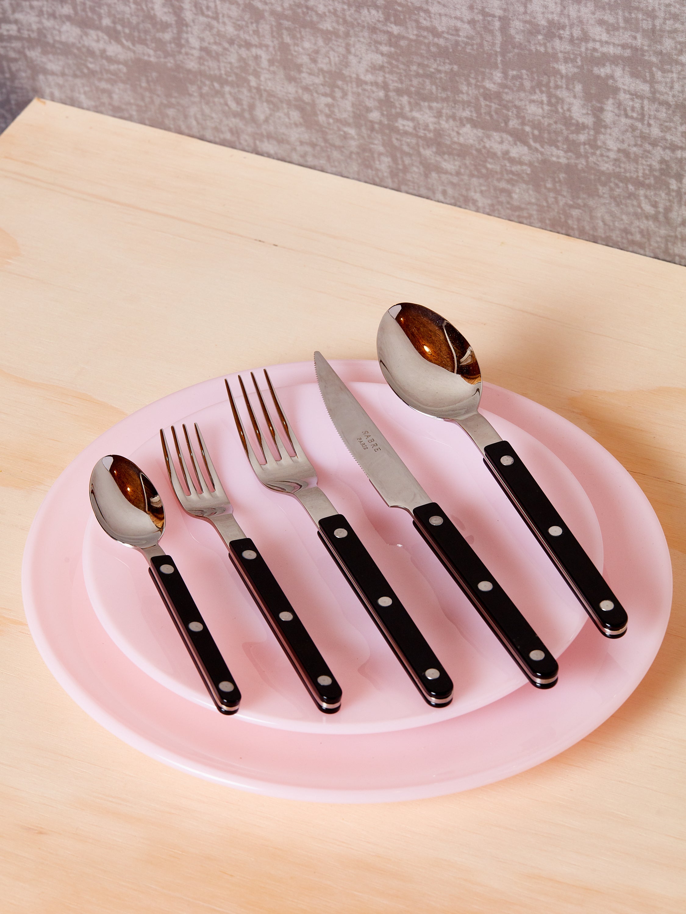 Five pieces of black bistro flatware sit atop a stack of two pink plates by Mosser.