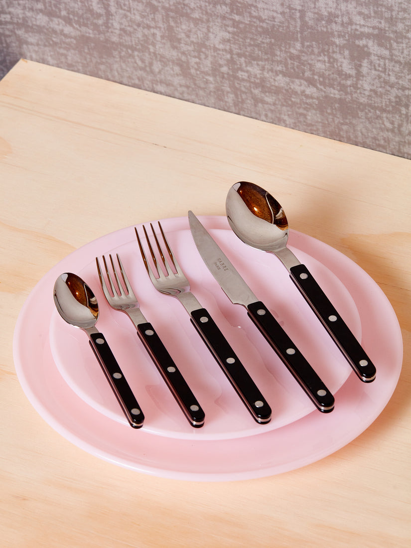 Five pieces of black bistro flatware sit atop a stack of two pink plates by Mosser.