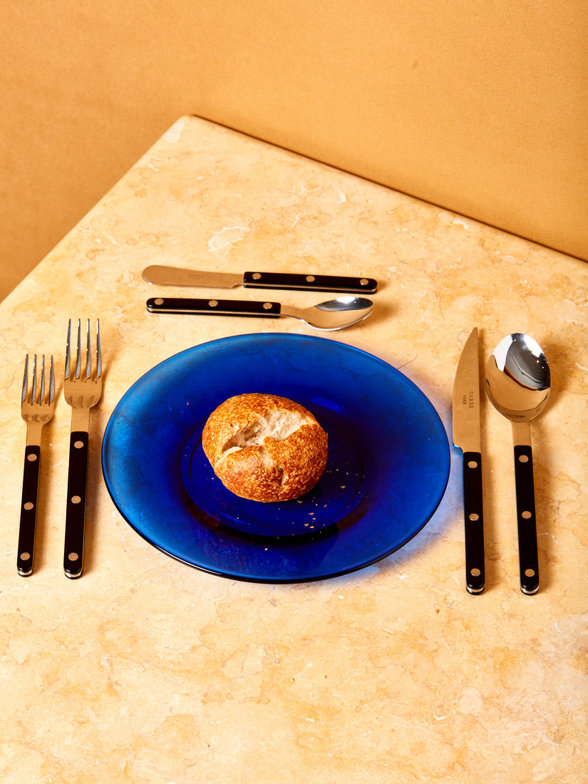 A small bread roll on a blue plate surrounded by a place setting of black bistro flatware by Sabre.
