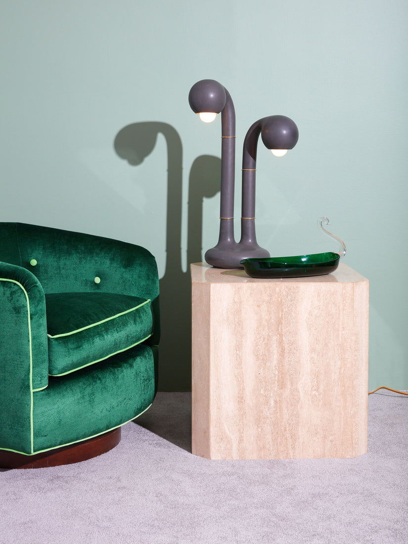 Entler's two globe matte charcoal table lamp sits atop a travertine side table next to a vintage glass dish. Sitting next to the table is an emerald green velvet club chair.