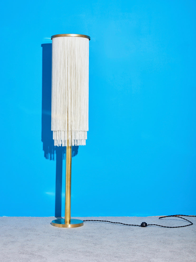 Barbara the Fringe Floor Lamp, a brass lamp with white silk fringe against a grey carpet and bright blue wall.