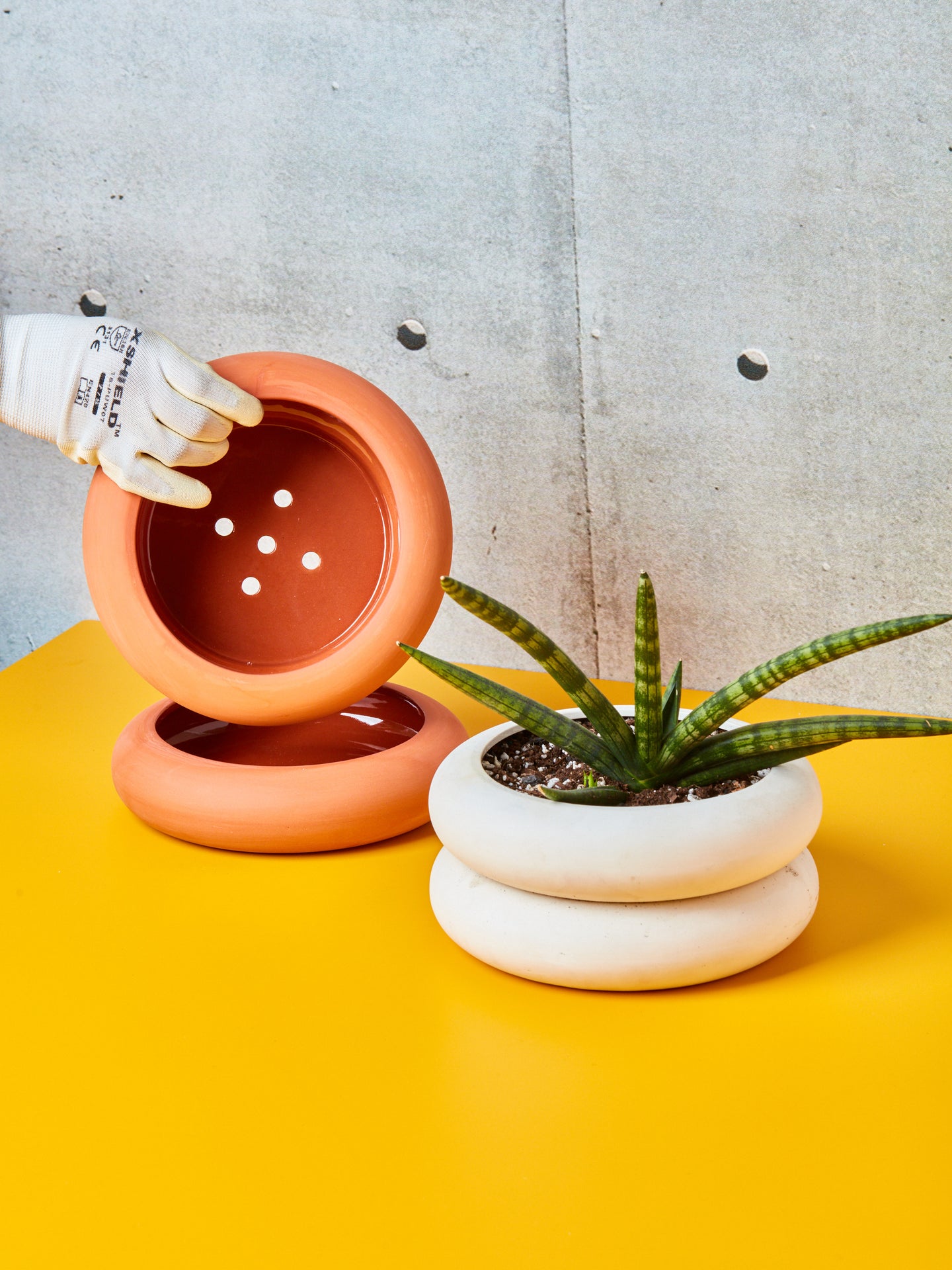 A hand lifts up the short terracotta stacking planter to reveal its drainage holes. A succulent is potted in the short white stacking plater sitting right.