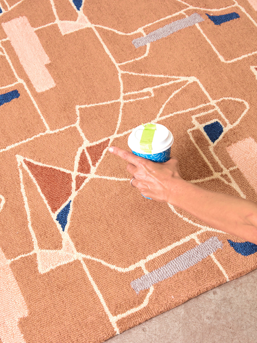 A hand holds a coffee cup above The Long Goodbye Rug.
