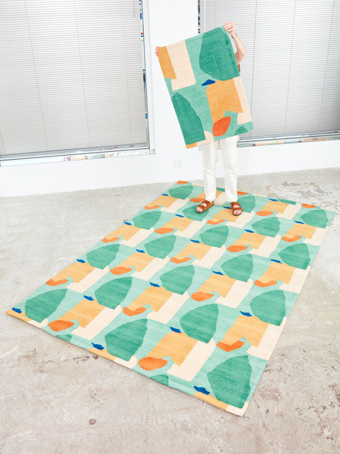 Someone holds up a 2x3 Summer Visitors Rug by Cold Picnic while standing at the back edge of a 4x6 version.