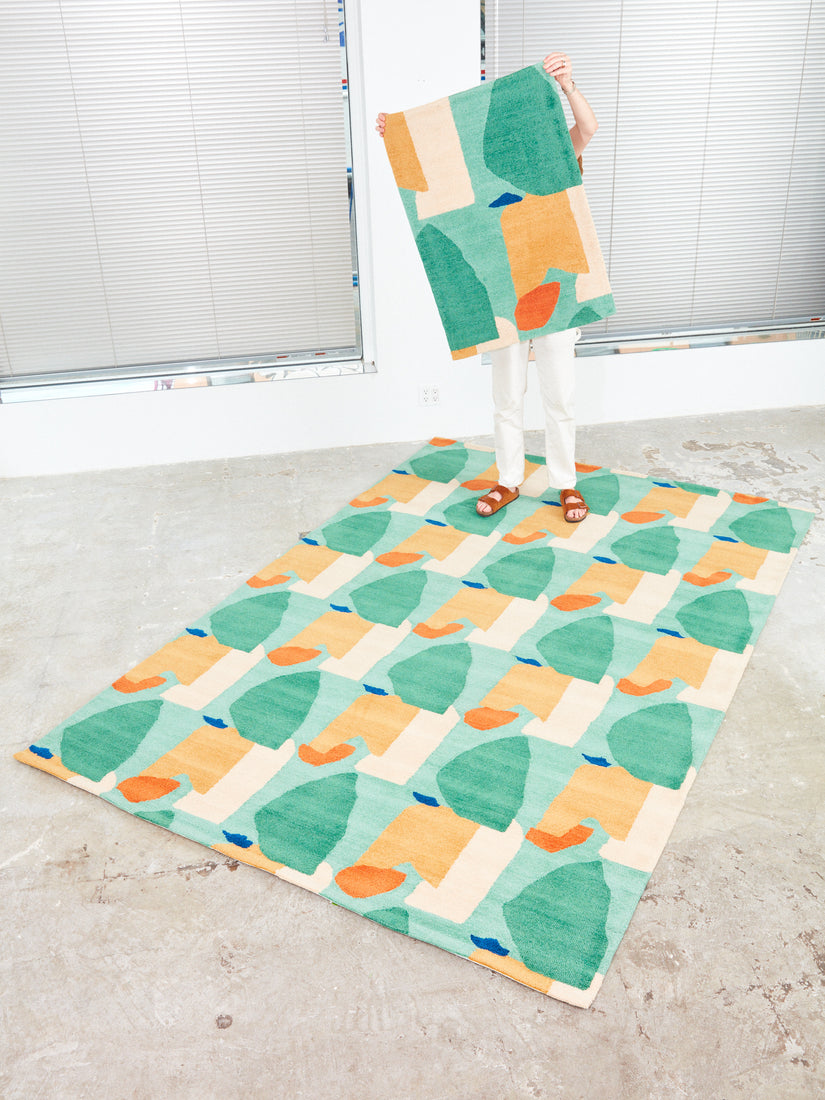 Someone holds up a 2x3 Summer Visitors Rug by Cold Picnic while standing at the back edge of a 4x6 version.