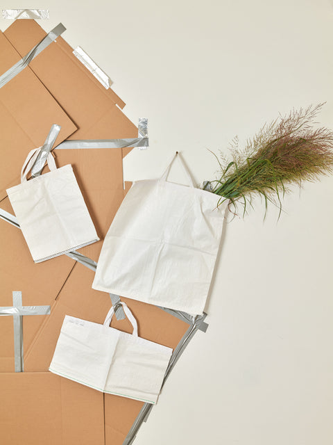 Tyvek tote bags of all different shapes hung on a wall of collaged cardboard. The largest bag is full of branches.