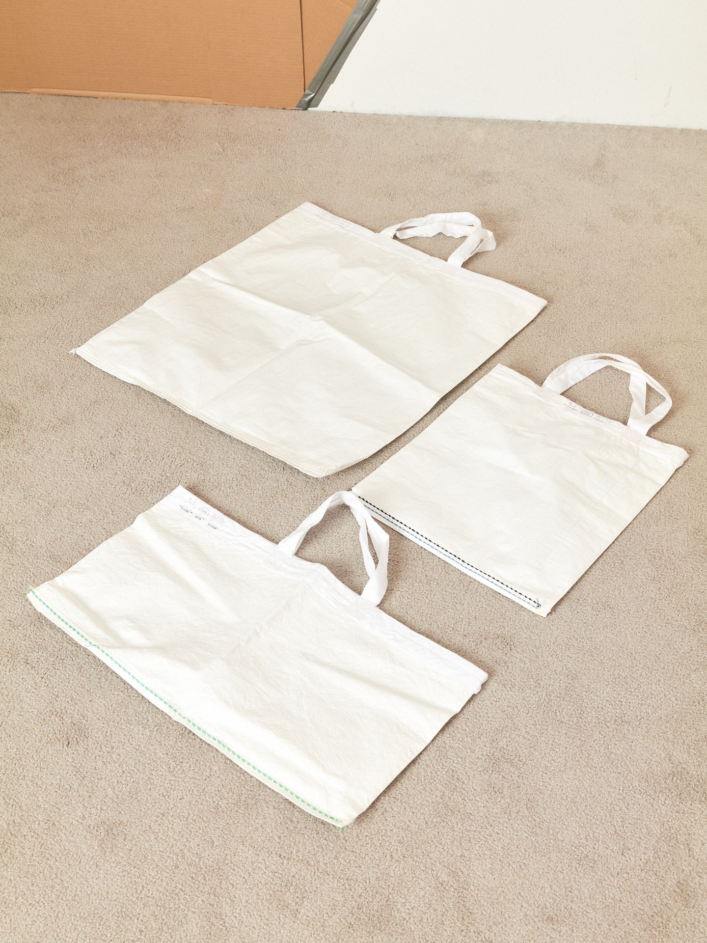 White tote bags in 3 different sizes laying neatly on a carpeted floor.