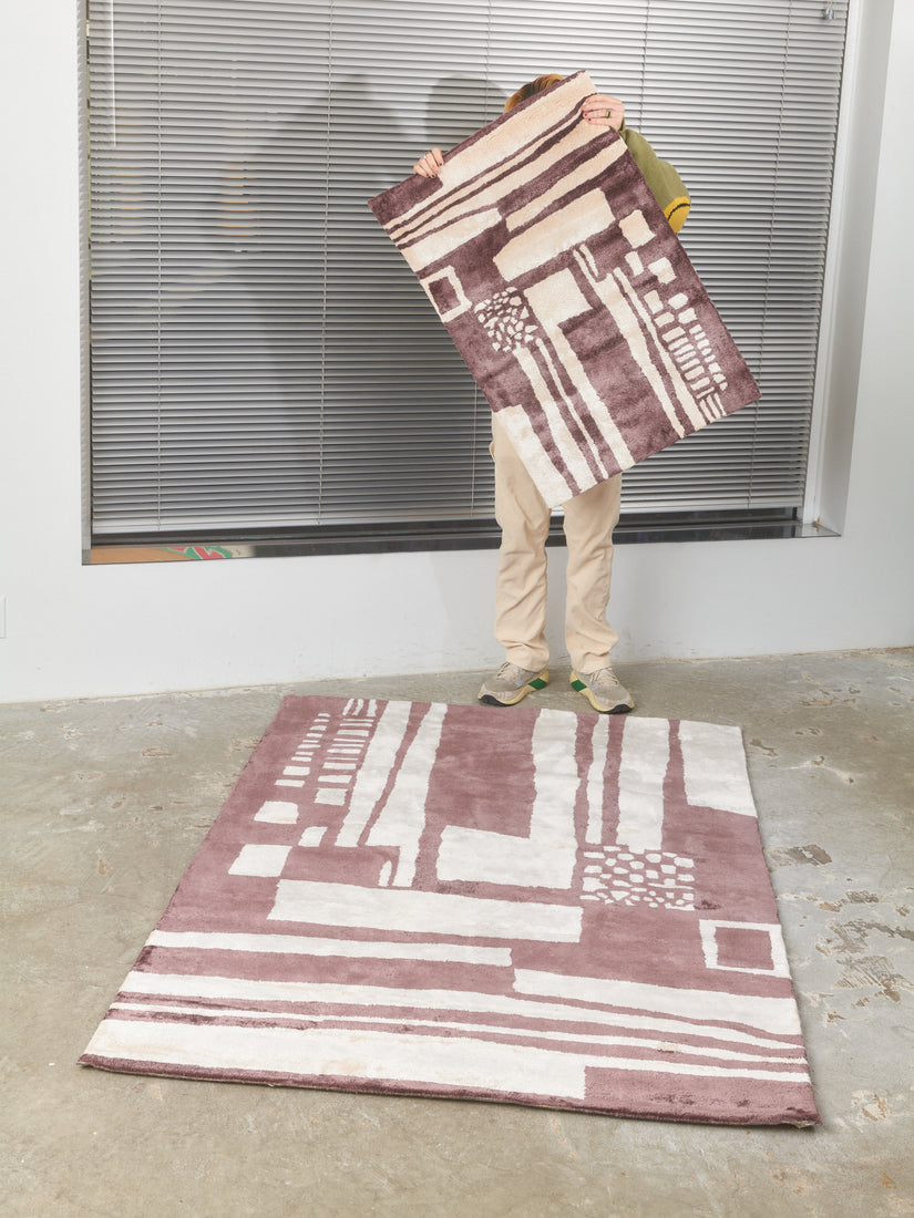 Battenburg Rug in Chocolate Vanilla by Cold Picnic features a dark charcoal and ivory abstract pattern. The subject in the photograph is holding up a 2ft by 3ft rug covering their upper body while standing along the edge of a 4ft by 6ft version of the same rug laying on a concrete floor.