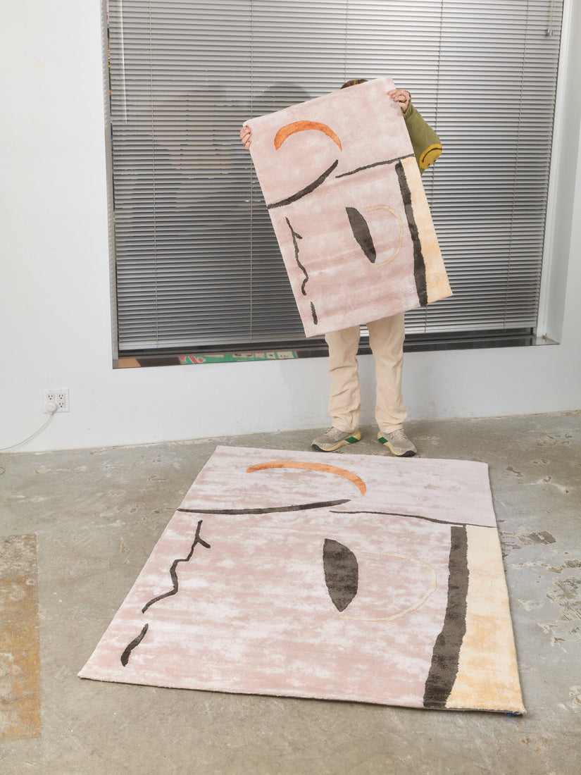 Someone holds up a 2x3 Taro Rug by Cold Picnic standing behind a 4x6 which lays on the concrete floor.
