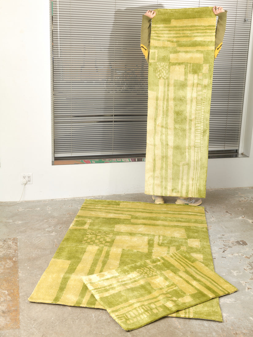 Someone holds up a 2x6 version of a Battenberg Rug in Matcha by Cold Picnic standing at the back edge of a 4x6 on the floor. Atop the 4x6 is a 2x3 version of the same rug.