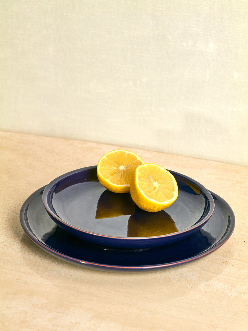 A blue salad plate with two halves of a lemon sits atop a blue dinner plate by Trame.