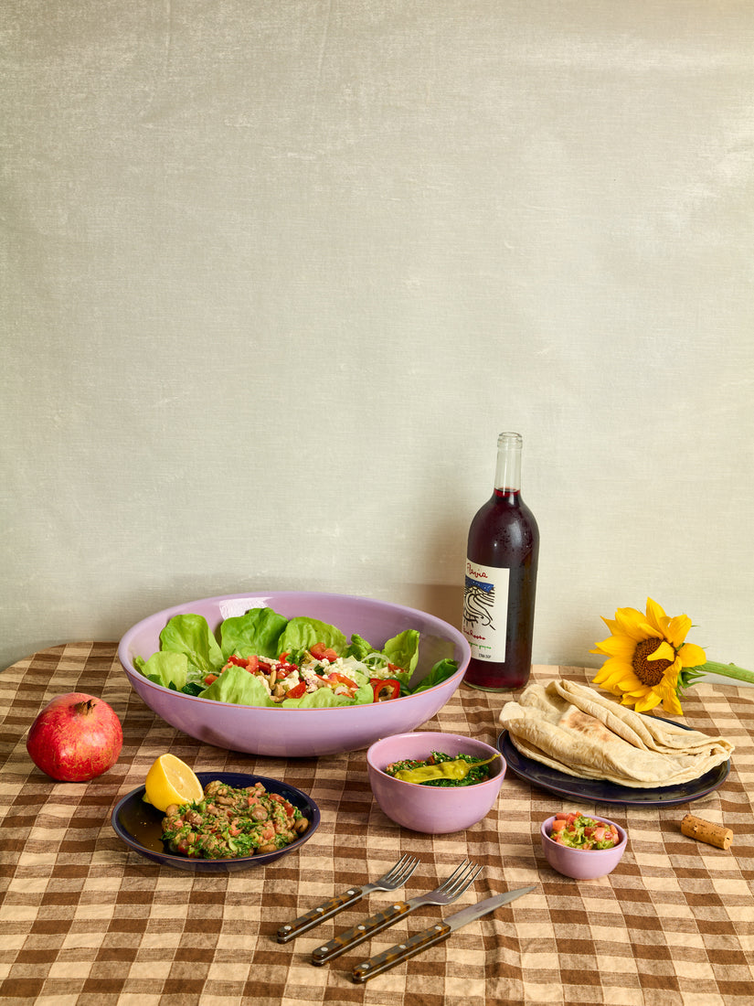 A full dinner spread on a brown gingham table cloth. A large salad, chickpeas, pita, a bottle of wine, a sunflower, a pomegranate, and smaller salads arranged in a still life of ceramic dinnerware by Trame. 