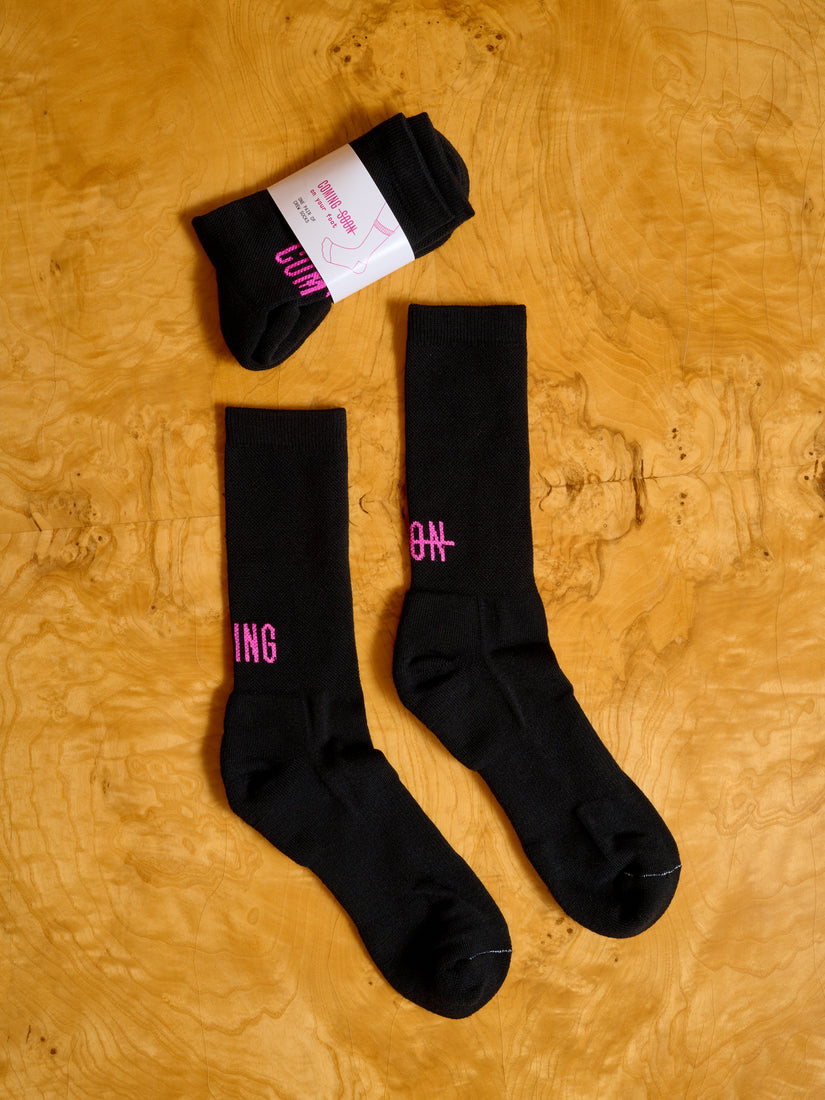 Two pairs of black Coming Soon socks, one in its packaging.
