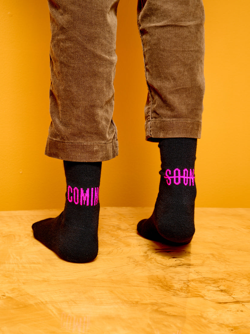 Someone wearing black Coming Soon socks with pink logo.