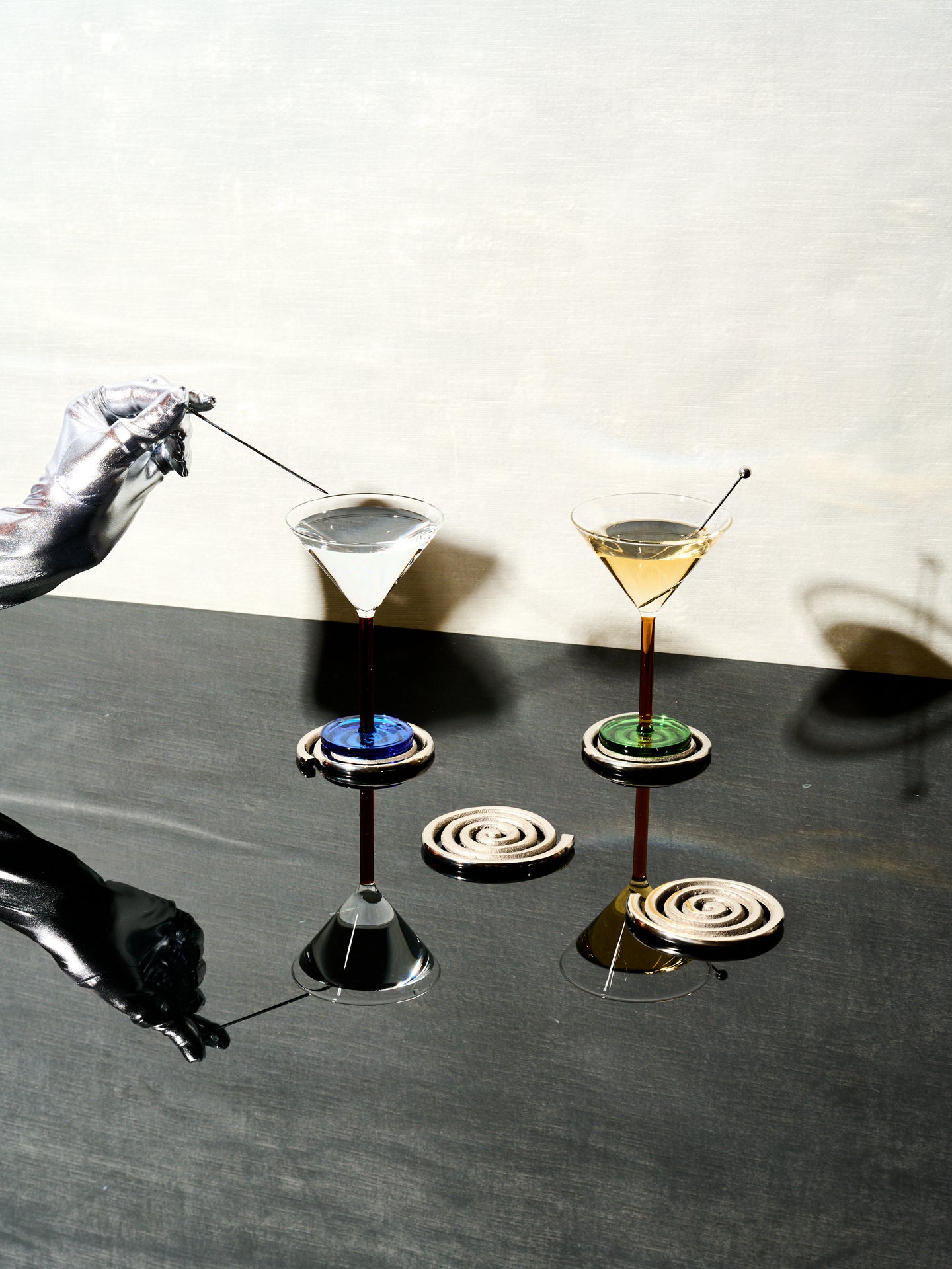 A silver hand pulls the skewer from a pair of Piano Cocktail Glasses that sit atop a set of Spiral Coasters by Sophie Lou Jacobsen.
