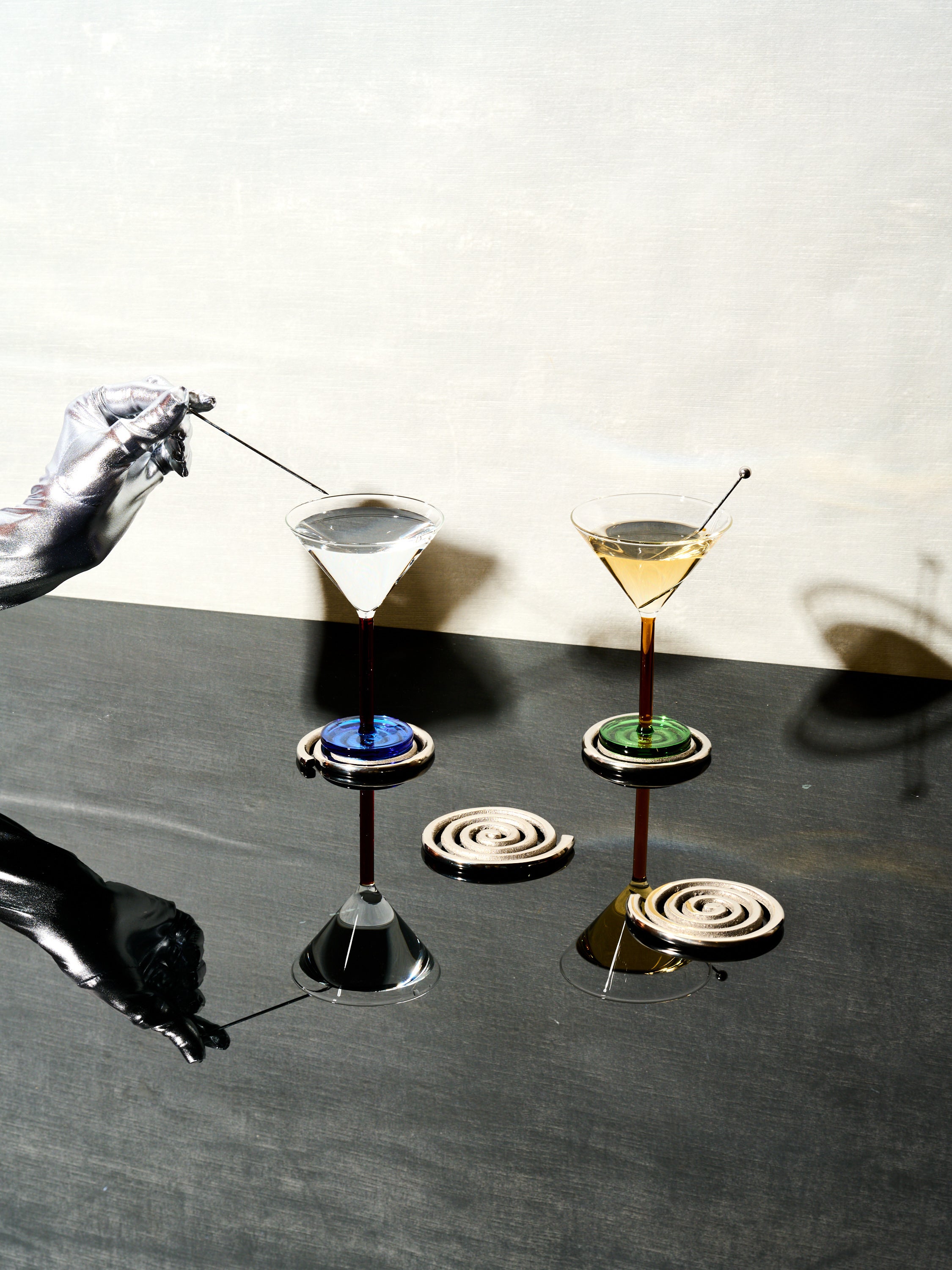 A silver hand pulls the skewer from a pair of Piano Cocktail Glasses that sit atop a set of Spiral Coasters by Sophie Lou Jacobsen.