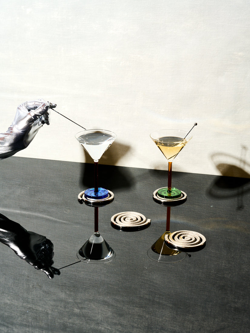 A silver hand pulls the skewer from a pair of Piano Cocktail Glasses that sit atop a set of Spiral Coasters by Sophie Lou Jacobsen.