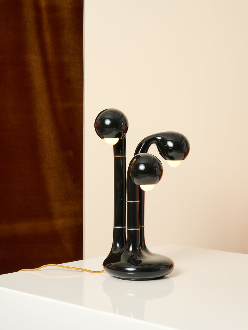 A glossy black three globe table lamp by Entler sitting on a white table against a beige wall and brown velvet curtain. The lamp features smooth curvature and brass hardware.