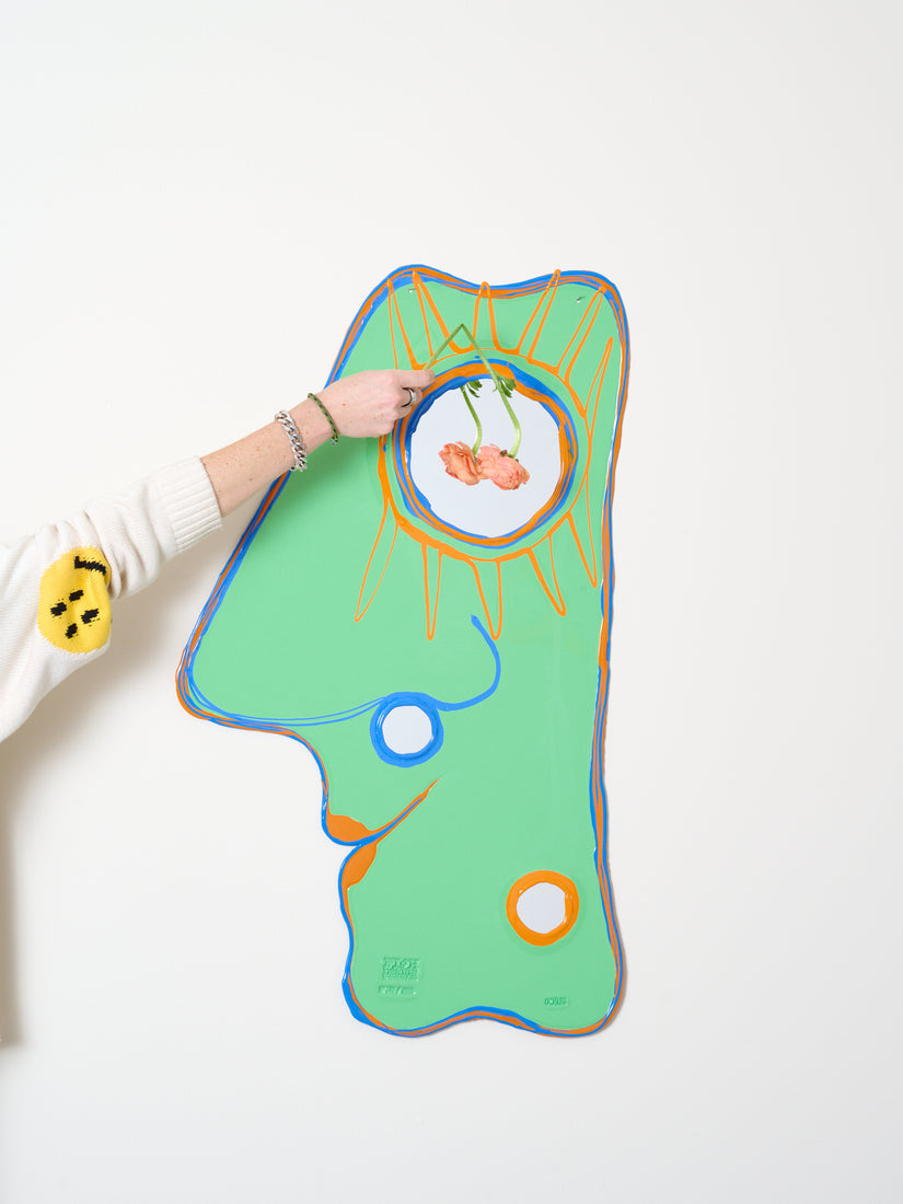 Abstract face shaped mirror with highlights of blue and orange. A hand holds up a limp flower in front of the larger mirror portion.