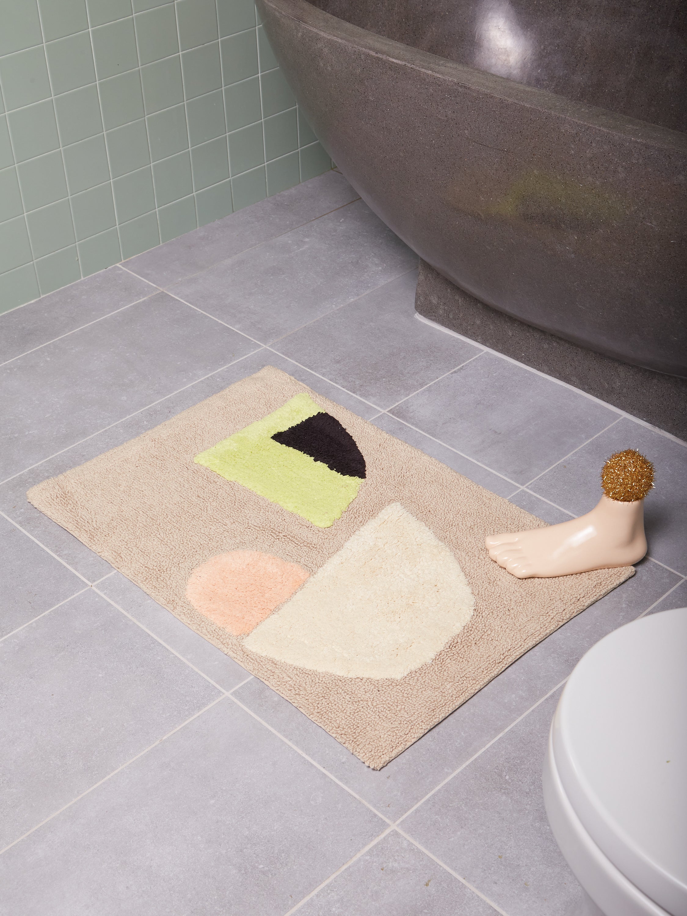 Eclipse Bath Mat in a tiled bathroom in front of a stone tub with a ceramic foot on the corner of the bath mat.