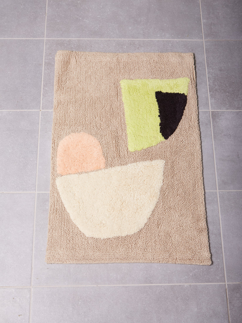 Eclipse Bath Mat by Cold Picnic against a grey tiled floor. Mostly beige mat with abstract forms of peach, cream, lime green, and black.