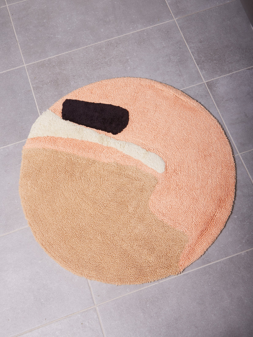 A round bath mat with abstract design that's mostly peach and beige with an accent of black and accent of white.