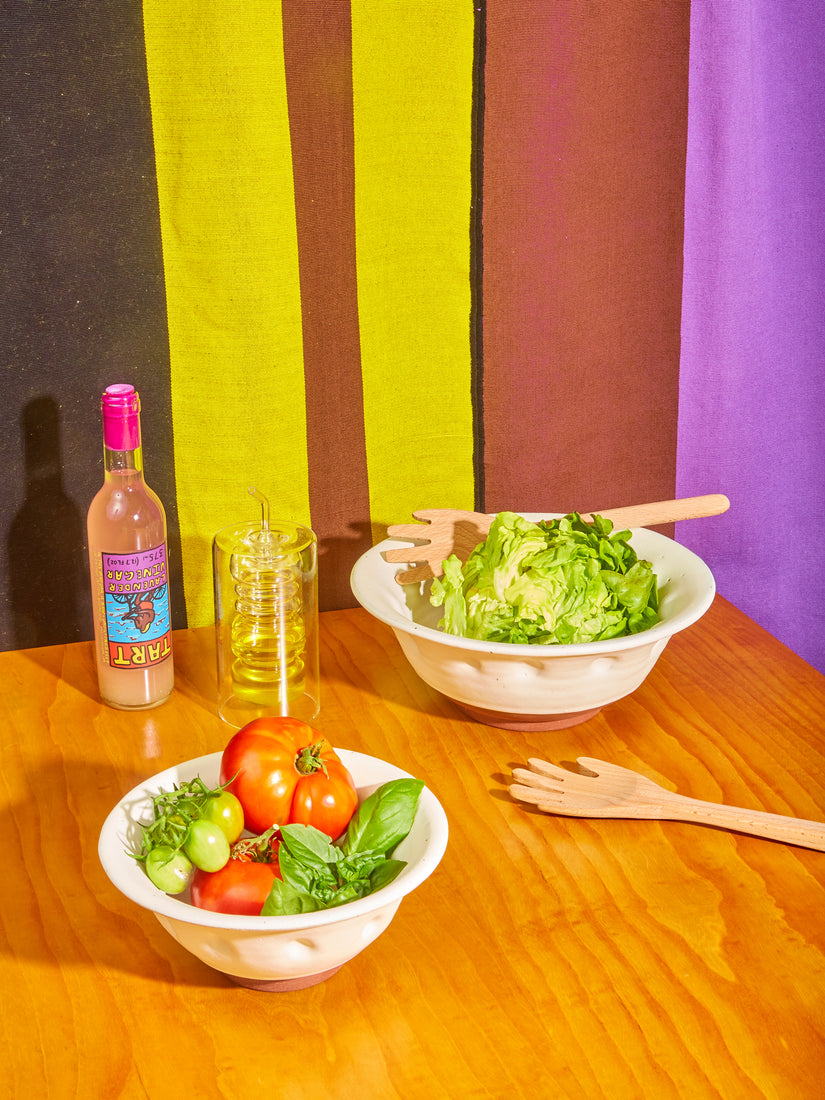 A tablescape of Dimpled Ceramic Serving Bowls by Valtierra Ceramica full of fresh vegetables, Hands Serving Friends by Areaware, Tart Vinegar, and a Rings Oil Bottle.
