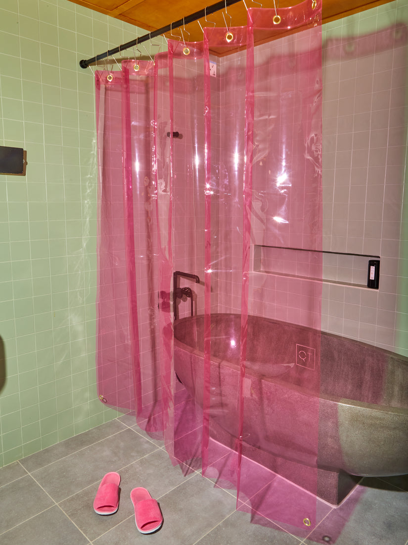 A transparent pink Sun Shower Curtain by Quiet Town in a sage teal tiled bathroom. Pink terrycloth slides sit in front of the shower curtain.